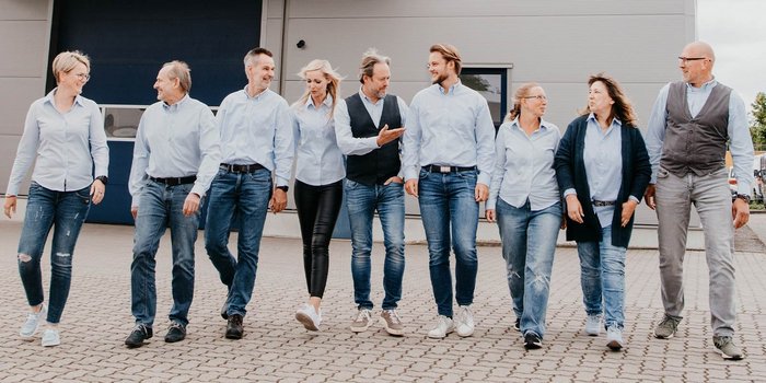 Eine Gruppe von neun Personen in blauen Hemden und Jeans geht lächelnd auf einem gepflasterten Weg entlang.