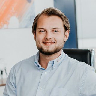 Der Mann trägt ein hellblaues Hemd und sitzt in einem Bürostuhl, während er in die Kamera lächelt, umgeben von Büromöbeln.