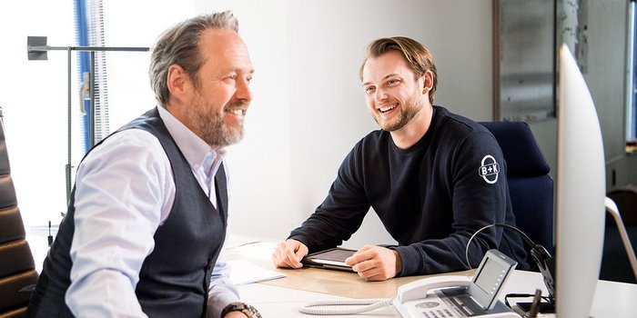 Ein älterer Mann in einem Anzug und ein jüngerer Mann in einem Sweatshirt unterhalten sich freundlich an einem Schreibtisch.