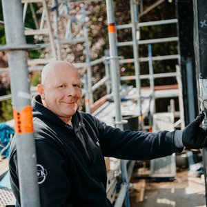 Der Bauarbeiter in einem schwarzen Hoodie zeigt ein freundliches Lächeln, während er an einem Gerüst steht und arbeitet.