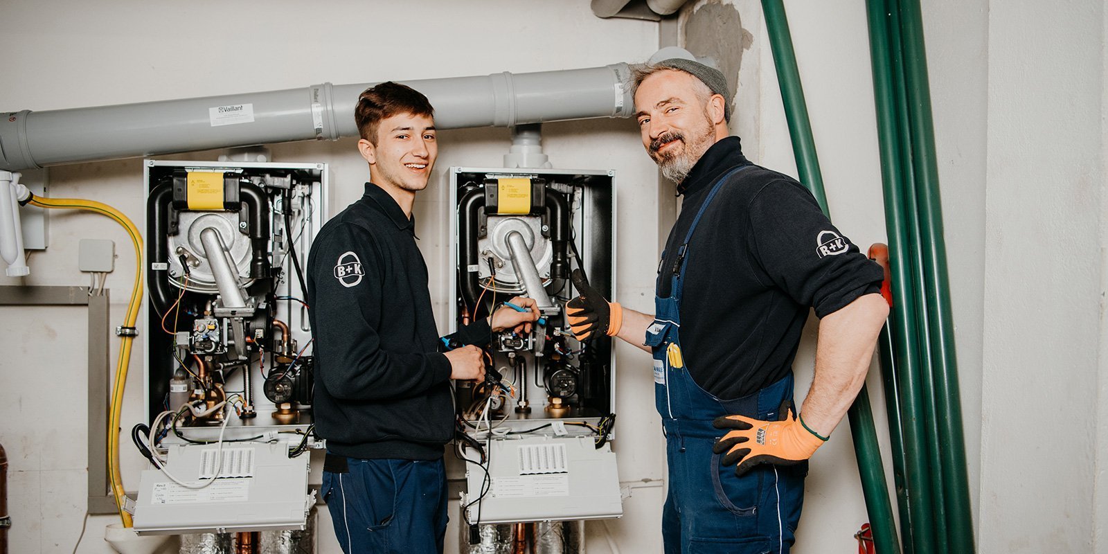 Ein junger und ein erfahrener Techniker stehen vor einer Heizungsanlage und zeigen Daumen hoch, beide tragen Handschuhe.
