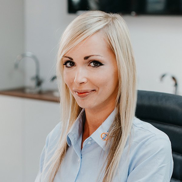 Eine blonde Frau mit langen Haaren lächelt freundlich in einem modernen Büro, während sie auf einem Stuhl sitzt.