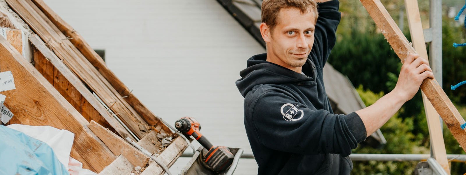 Ein Handwerker hebt ein Holzstück an, während er auf einem Dach arbeitet, mit einem Lächeln im Gesicht und einem Bohrer in der Nähe.