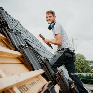 Auf dem Dach eines Hauses installiert ein Handwerker mit einem freundlichen Lächeln Dachziegel, umgeben von Wolken und Bäumen.