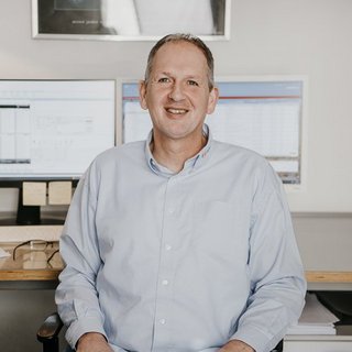 Der Mann wirkt freundlich und entspannt, während er in einem modernen Büro mit technischen Geräten umgeben ist.