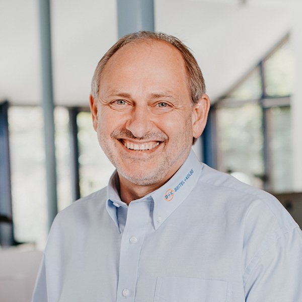 Der Mann mit dem freundlichen Lächeln hat ein hellblaues Hemd an und posiert in einem lichtdurchfluteten Raum mit Glaswänden.