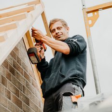 Auf einem Gerüst arbeitet ein Handwerker mit einem Akkuschrauber, um eine Dachkonstruktion zu montieren, und wirkt dabei zufrieden.