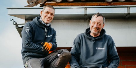 Die beiden Männer auf dem Dach zeigen Freude, während sie in Arbeitskleidung und Handschuhen posieren.
