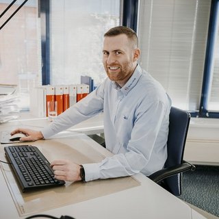 Ein freundlicher Mann mit Bart sitzt an einem Schreibtisch, umgeben von Aktenordnern und einem großen Fenster.