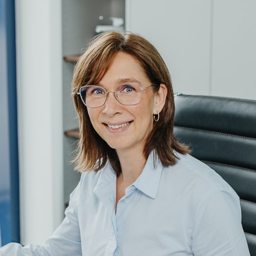 Die Frau mit Brille hat eine positive Ausstrahlung und sitzt an einem Schreibtisch, der mit modernen Büroelementen ausgestattet ist.