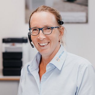 Die Frau trägt ein hellblaues Hemd und ein Headset, während sie in einem hellen Büro mit einem warmen Lächeln posiert.