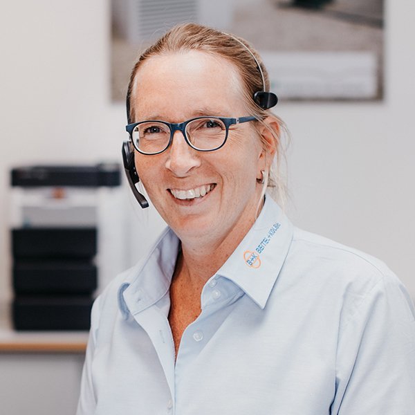 Die Frau trägt ein hellblaues Hemd und ein Headset, während sie in einem hellen Büro mit einem warmen Lächeln posiert.