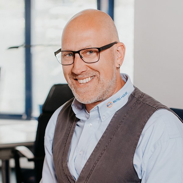 Ein lächelnder Mann mit Brille trägt ein Hemd und eine Weste, während er in einem modernen Büro sitzt.