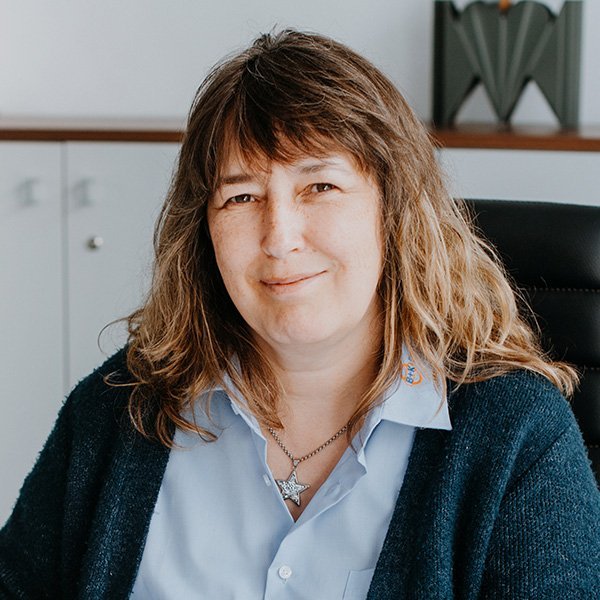 In einem modernen Büro lächelt eine Frau mit einem blauen Hemd und einem Sternanhänger, während sie auf einem Stuhl sitzt.