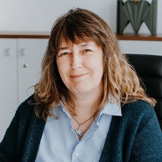 In einem modernen Büro lächelt eine Frau mit einem blauen Hemd und einem Sternanhänger, während sie auf einem Stuhl sitzt.