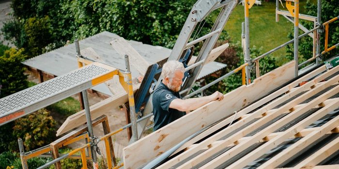 Auf einem Gerüst steht ein Handwerker, der Holzplatten auf einem Dach anbringt, während die Sonne scheint und die Natur blüht.
