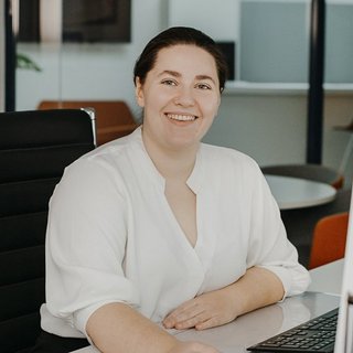 Die Frau trägt ein weißes Hemd und hat eine freundliche Ausstrahlung, während sie an ihrem Arbeitsplatz sitzt.