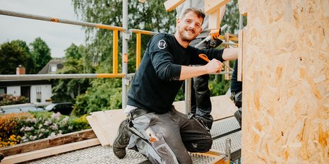 Auf einem Gerüst arbeitet ein Handwerker mit einem Werkzeug in der Hand, während im Hintergrund ein blühender Garten sichtbar ist.