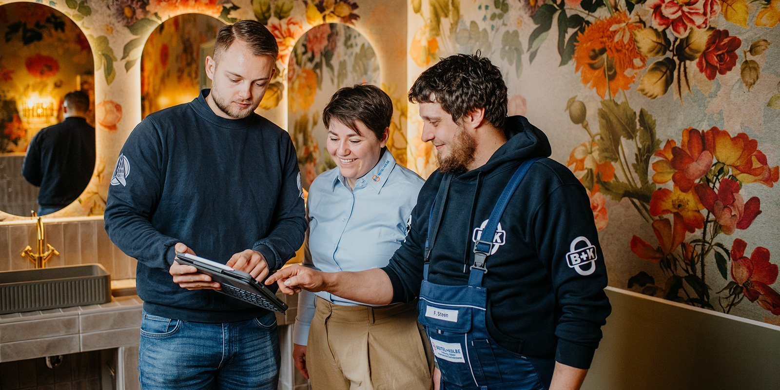Ein Mann zeigt auf ein Tablet, während eine Frau und ein weiterer Mann interessiert zuschauen, umgeben von bunten Blumenmotiven.