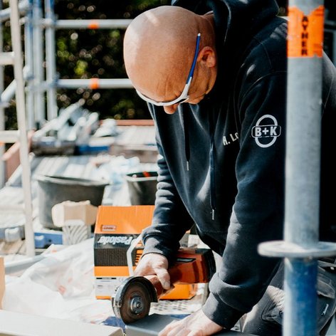 Der Handwerker bearbeitet Materialien auf einer Baustelle, umgeben von Werkzeugen und Baustellenzubehör, mit einem fokussierten Ausdruck.
