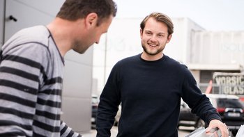 Ein lächelnder Mann interagiert mit einem anderen, der sich auf eine Aufgabe konzentriert, umgeben von einem modernen Gebäude.