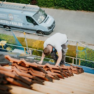 Ein Dachdecker beugt sich über die Ziegel, die er verlegt, während ein blauer Lieferwagen im Hintergrund sichtbar ist.