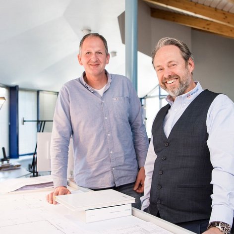 Ein modernes Büro mit großen Fenstern zeigt zwei Männer, die an einem Tisch mit Architekturplänen stehen und freundlich in die Kamera lächeln.