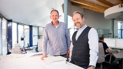 Ein modernes Büro mit großen Fenstern zeigt zwei Männer, die an einem Tisch mit Architekturplänen stehen und freundlich in die Kamera lächeln.