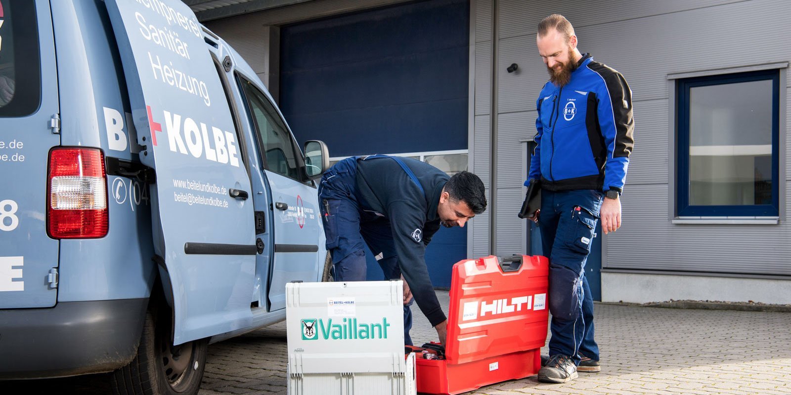 Vor einem Lieferwagen arbeiten zwei Männer, einer überprüft Werkzeuge in einer roten Kiste, der andere beobachtet.