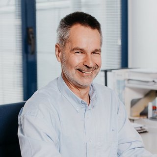 Der Mann trägt ein hellblaues Hemd und hat einen freundlichen Ausdruck, während er in einem modernen Büro sitzt.