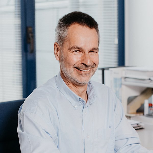 Der Mann trägt ein hellblaues Hemd und hat einen freundlichen Ausdruck, während er in einem modernen Büro sitzt.