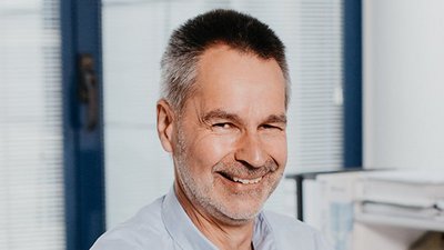 Der Mann trägt ein hellblaues Hemd und hat einen freundlichen Ausdruck, während er in einem modernen Büro sitzt.